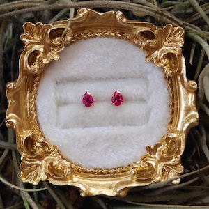 May include: A pair of teardrop-shaped, vibrant pink stud earrings set in gold, displayed within a small, ornate, metallic gold-colored frame featuring a white, plush, velvet-like interior lining with two horizontal slots.