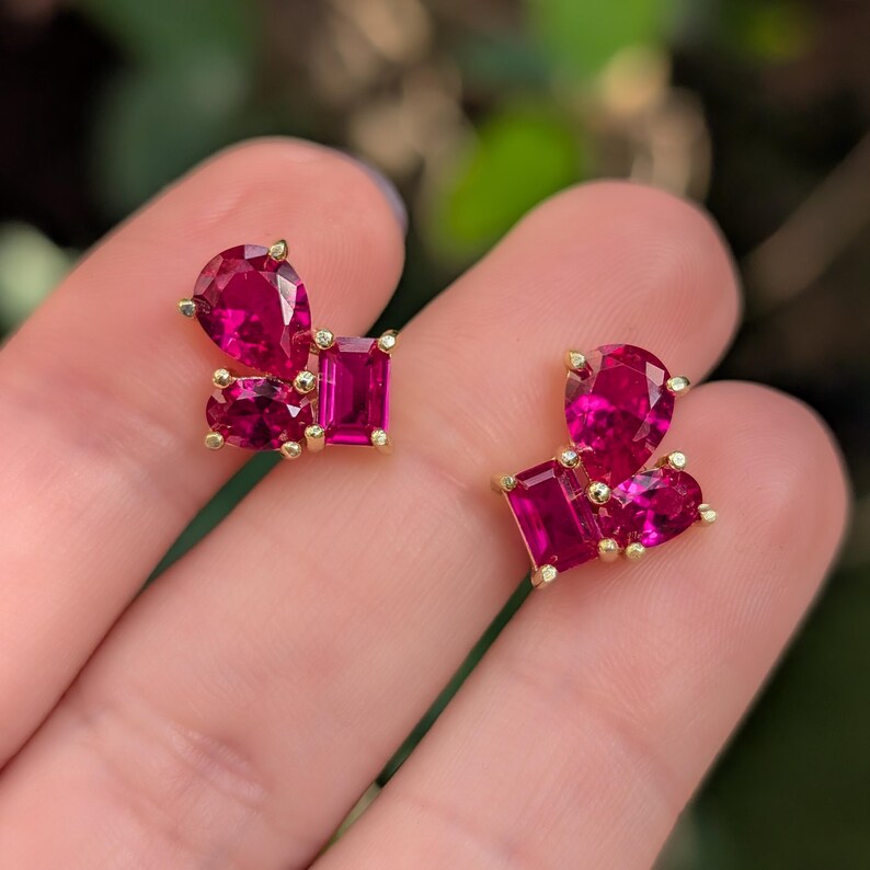 Peut inclure: Une paire de boucles d'oreilles en or avec des grappes de pierres pr&eacute;cieuses rouge rubis vibrantes. Chaque boucle d'oreille pr&eacute;sente un arrangement unique de pierres en forme de poire, rectangulaire et en c&oelig;ur, cr&eacute;ant un design &eacute;blouissant et accrocheur.