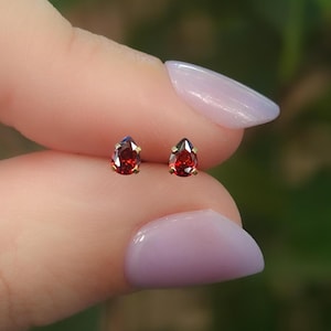 May include: A pair of stud earrings featuring pear-shaped, deep red gemstones set in gold-toned metal prongs. The teardrop-shaped stones are faceted, reflecting light, and are held securely by small metal claws on each individual earring.