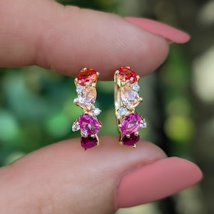 May include: A pair of gold hoop earrings featuring a gradient of multi-colored gemstones. Each hoop is adorned with a sequence of oval and pear-shaped stones in shades of orange, peach, pink, and deep red, accented by small clear stones.
