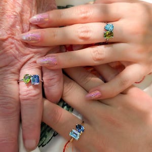 May include: Three rings featuring dual-stone designs on thin metallic bands. Each ring showcases a combination of two distinct gemstones in various shapes, including pear, emerald, and square cuts, in vibrant shades of blue, green, and light purple.
