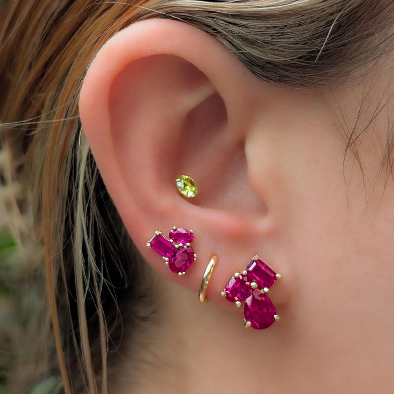 Peut inclure: Gros plan sur une oreille orn&eacute;e de plusieurs boucles d'oreilles. Un petit clou rond vert clair est en haut. En dessous, un anneau dor&eacute; et deux grappes de boucles d'oreilles dor&eacute;es avec des pierres pr&eacute;cieuses roses vives sont visibles, mettant en valeur un style de bijoux moderne.
