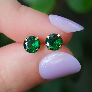 May include: A pair of round, faceted emerald green stud earrings set in gold-toned metal prongs. Each earring features a circular, vibrant green gemstone with a brilliant cut, held securely by four small metal claws in a classic stud design.