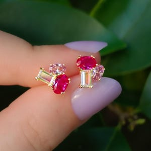 May include: A pair of gold stud earrings, each featuring a cluster of three gemstones: a vibrant red oval stone, a pale pink oval stone, and a clear rectangular baguette-cut stone, all set in a polished gold-tone metal base.