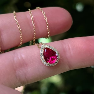 May include: A gold necklace featuring a pear-shaped, vibrant red gemstone pendant. The pendant is surrounded by a halo of small, clear gemstones. The chain is delicate and gold-colored, creating an elegant jewelry piece.