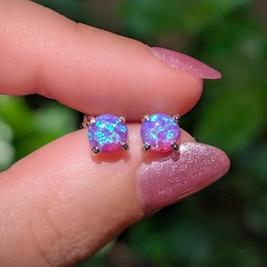 May include: A pair of round stud earrings featuring vibrant purple and blue iridescent opal-like stones set in silver-toned metal prongs. The circular stones display a speckled, reflective surface with bright flashes of light blue and violet hues.