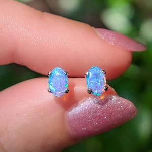 May include: A pair of oval-shaped stud earrings featuring iridescent blue opals set in silver-toned metal prongs. The stones display a vibrant play of color with flashes of bright cyan and light purple throughout the smooth, polished surfaces.