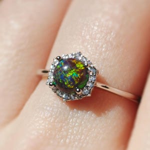 May include: A silver ring featuring a round, iridescent black opal gemstone surrounded by small, clear accent stones. The opal displays flashes of green, blue, and yellow. The ring is set on a thin, polished silver band.