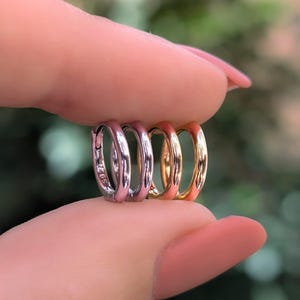 May include: Two small hoop earrings, one in a silver tone and one in a gold tone, held between fingers. The silver hoop features a visible S925 hallmark stamp on its side. Both earrings have a smooth, polished, circular metal design.