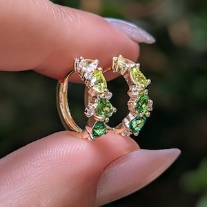 May include: A pair of gold hoop earrings featuring a gradient of pear-cut green gemstones and small clear accent stones. The stones transition from light lime green at the top to a deep emerald green at the bottom, set in a polished gold band.