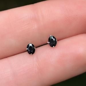 May include: A pair of stud earrings featuring oval-cut black gemstones set in silver-toned metal prongs. Each earring displays a faceted surface that reflects light, showcasing a deep, dark color profile and a classic, symmetrical oval shape.