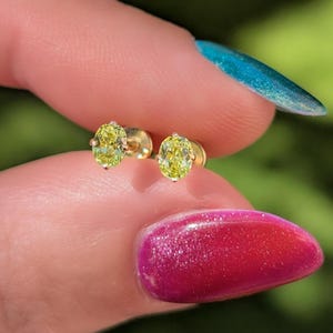 May include: A pair of gold stud earrings, each featuring an oval-shaped, light yellow gemstone. The earrings are held between fingers with brightly colored nail polish in the background. The earrings are a jewelry item.