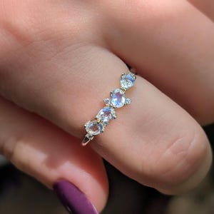 以下が含まれることがあります： 4つの楕円形で虹色に輝くムーンストーンが並んだシルバー調のリング。石は柔らかな青い光を放ち、それぞれのムーンストーンの間に小さな透明なアクセントストーンが配置された繊細な金属製のバンドにセットされています。