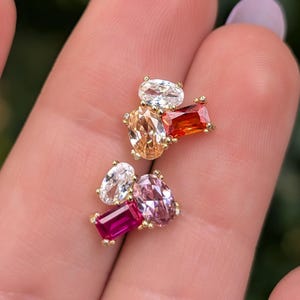May include: Close-up of two gold-toned stud earrings, each featuring a cluster of colorful gemstones. The gemstones include oval, rectangular, and pear shapes in shades of white, orange, red, pink, and magenta. The earrings are set against a blurred background.