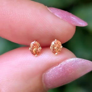 May include: A pair of stud earrings featuring oval-cut orange gemstones set in gold metal prongs. Each earring displays a faceted surface that reflects light, showcasing a warm, vibrant orange hue. The stones are secured by small metal claws.