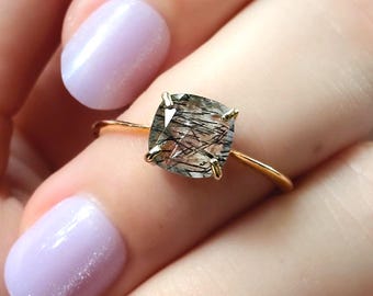 Può includere: Un anello in oro con una pietra preziosa di quarzo rutilato a taglio cuscino con visibili inclusioni nere simili ad aghi. La pietra trasparente è fissata in una montatura a quattro griffe su una sottile fascia in oro lucido.