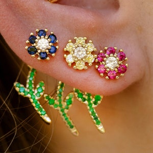 May include: Three gold flower earrings with colorful gemstones. The earrings are in the shape of a flower with a center diamond and surrounding gemstones. The flowers are blue, yellow, and red.