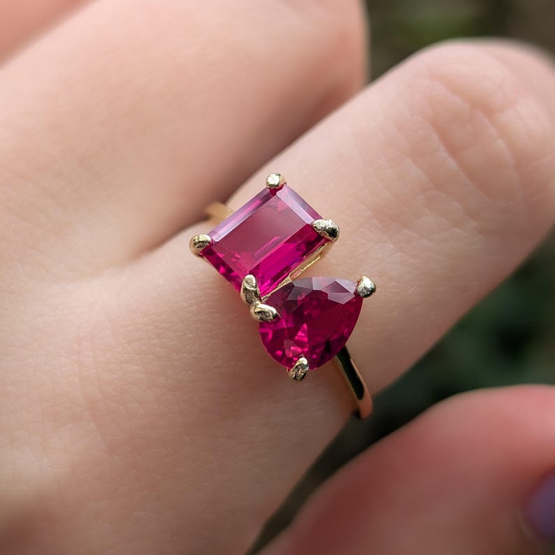 May include: A gold ring with two red-pink gemstones. One is rectangular, the other heart-shaped. The ring's band is thin, polished gold, and the gemstones are held by small gold prongs. The ring is a piece of jewellery.