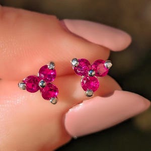 May include: A pair of stud earrings featuring a three-stone cluster design. Each earring consists of three round, vibrant pink-red gemstones arranged in a triangular clover shape, set in a polished silver-toned metal setting with small prongs.