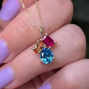 May include: A gold-colored necklace featuring a cluster of colorful gemstones. The pendant includes a pear-shaped blue gem, a rectangular pink gem, and two smaller orange and yellow gems. The necklace is displayed against a blurred background.
