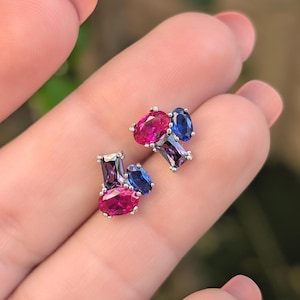 May include: A pair of stud earrings featuring a cluster design. Each earring has a silver setting and is adorned with oval and rectangular gemstones in shades of pink, purple, and blue.