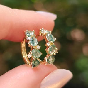 May include: A pair of gold hoop earrings featuring multiple round and pear-shaped green moss agate gemstones interspersed with small, clear, round-cut white stones set along the front-facing curve of each hoop.
