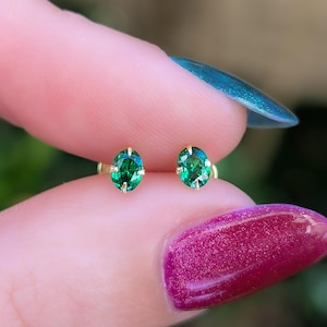 Puede incluir: Un par de pendientes de oro con tachuelas, cada uno con una piedra preciosa verde ovalada. Las piedras están engastadas en un engaste de cuatro puntas, mostrando su color y brillo. Los pendientes se muestran sobre un fondo borroso.