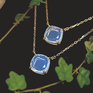 May include: Two delicate necklaces with square, light blue gemstone pendants. The gemstones are set in a gold-colored metal and hang from fine chains. Green leaves and vines frame the jewelry against a dark background.
