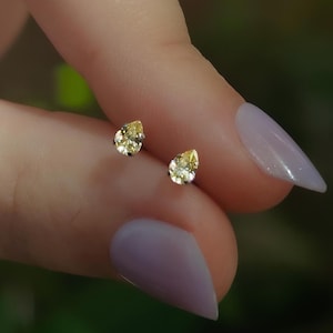 May include: A pair of stud earrings featuring pear-shaped yellow gemstones set in silver-toned metal. Each earring has a faceted surface that reflects light, showcasing a clear, pale yellow hue in a classic teardrop silhouette.