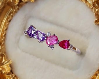 Puede incluir: Un anillo de tono plateado con una hilera de cuatro piedras preciosas multicolores en cortes de pera, óvalo y triángulo, en tonos morado, rosa y rojo, acentuadas por pequeñas piedras claras en una banda metálica fina.