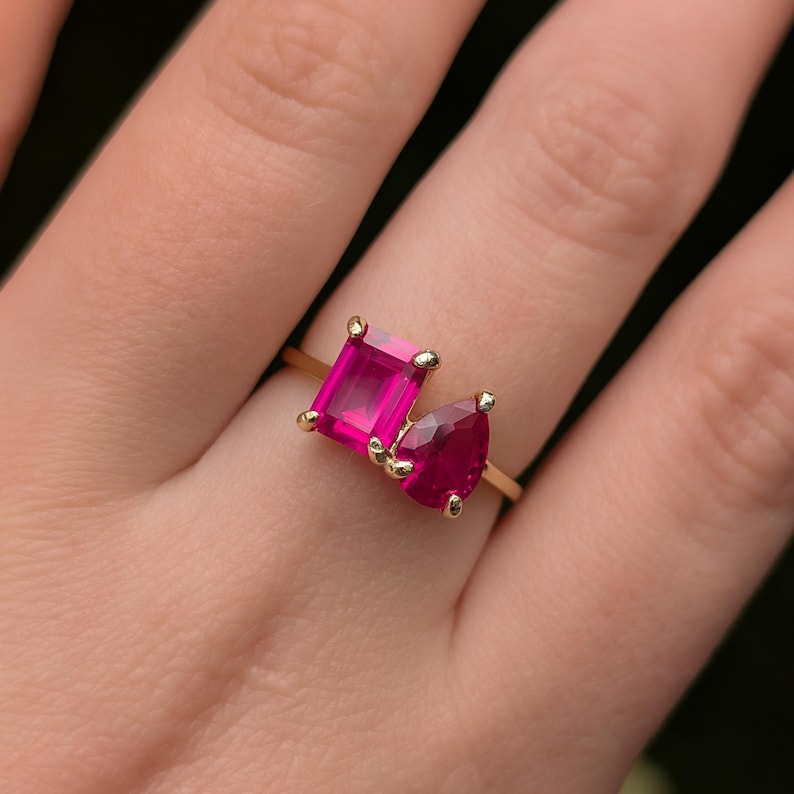 May include: A gold ring with two bright pink gemstones, one emerald cut and one pear cut. The ring has a slender, polished gold band, and the gemstones are set close together. The gemstones are a vivid shade of pink, creating a striking contrast against the gold.