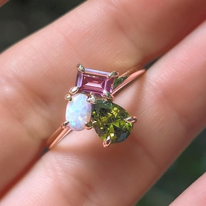 May include: A rose gold ring featuring a square pink gemstone, an oval white gemstone, and a pear-shaped green gemstone. The ring is set against a blurred background.