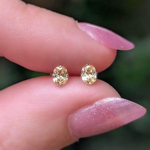 May include: A pair of stud earrings featuring oval-cut champagne-colored gemstones set in silver-toned metal prongs. Each earring displays a faceted surface that reflects light, showcasing a warm, golden-brown hue in a classic, simple design.