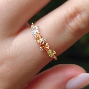 Op de afbeelding: Een gouden ring met een cluster van peervormige edelstenen in de kleuren transparant, perzik, oranje en geel, geaccentueerd door kleine ronde heldere stenen gezet in een dunne, gepolijste gouden band die om de vinger zit.