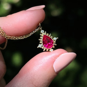 May include: A gold necklace with a pendant featuring a vibrant, teardrop-shaped, red gemstone. The pendant is surrounded by smaller, clear gemstones and a gold filigree design. The chain is a delicate gold link. The background is a blurred green.