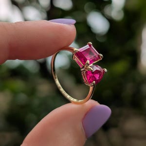 May include: A gold ring with two bright pink gemstones, one square and one heart-shaped. The ring has a slender gold band, and the gemstones are set close together. The design is striking and would make a lovely gift.