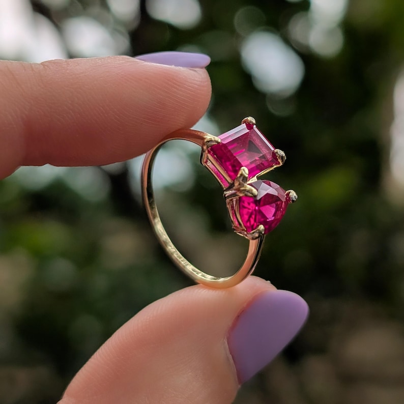 May include: A gold ring with two bright pink gemstones, one square and one heart-shaped. The ring has a slender gold band, and the gemstones are set close together. The design is striking and would make a lovely gift.