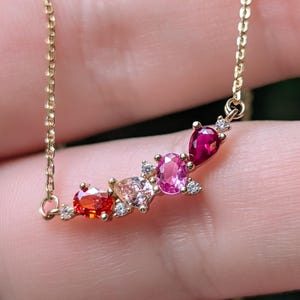 May include: A gold chain necklace featuring a curved pendant set with a gradient of multi-colored gemstones including a red oval, a peach pear-shape, a pink oval, and a deep red pear-shape stone, accented by small clear round white diamonds.