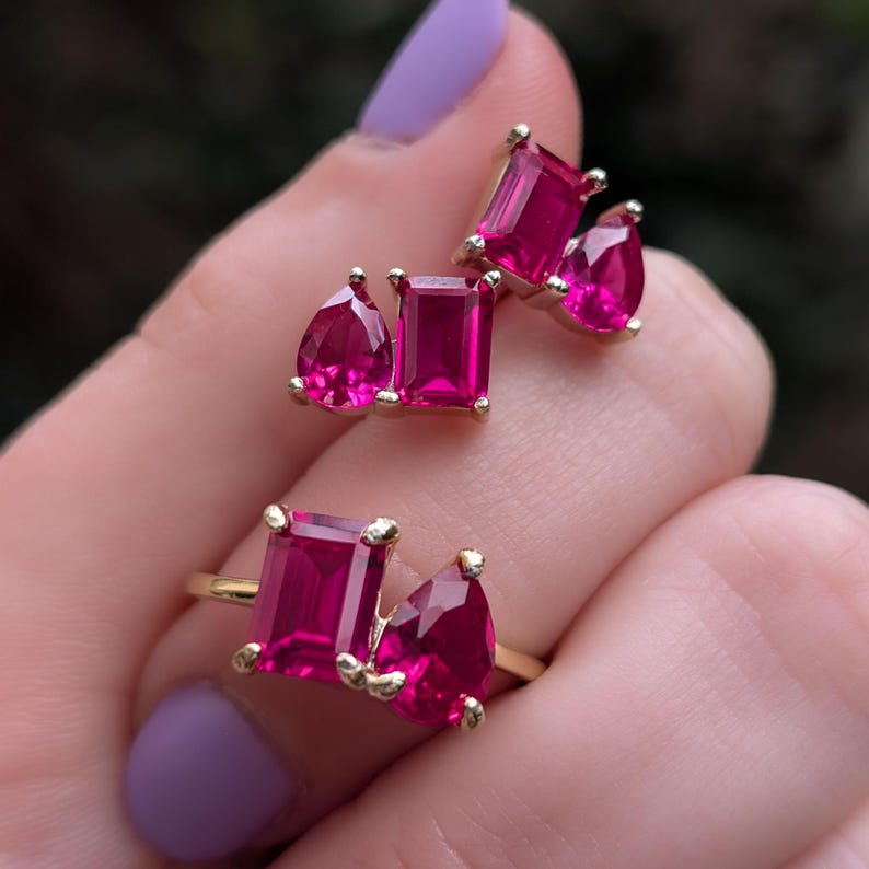 May include: Gold-toned jewellery set with bright pink gemstones. The set includes a ring and a pair of earrings, each featuring rectangular and pear-shaped stones. The gemstones are set in a gold metal setting.