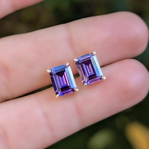 May include: A pair of emerald-cut stud earrings with a gold setting. The earrings feature a vibrant purple and blue gemstone. The earrings are displayed on a person's hand.