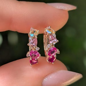 Puede incluir: Un par de pendientes de aro pequeños hechos de un material metálico, cada uno con un grupo de piedras preciosas de varias formas en tonos rosa, magenta y blanco iridiscente, dispuestos a lo largo de la curva frontal del diseño circular.
