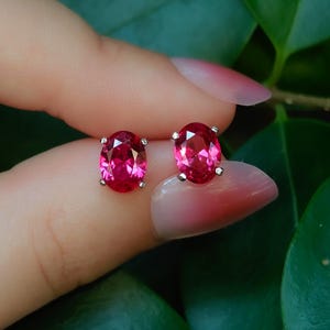 May include: A pair of oval-cut, vibrant pinkish-red gemstone stud earrings. Each stone is secured in a four-prong silver-toned metal setting, showcasing a faceted surface that reflects light with a brilliant, clear, and deep saturated color.