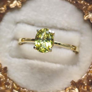 May include: A gold ring featuring a central oval-cut, lime-green gemstone held by four prongs. The band is thin and polished gold, showcasing a faceted stone that reflects light with a bright, yellowish-green hue set against a soft, neutral background.
