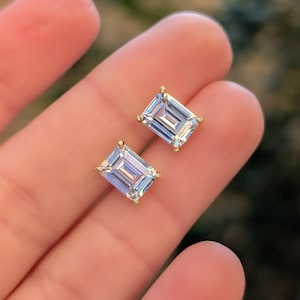 May include: Two emerald-cut, clear gemstone stud earrings with gold-coloured settings. The earrings are rectangular with multiple facets, reflecting light. The gold settings have small, decorative prongs. The earrings are displayed against a blurred background.