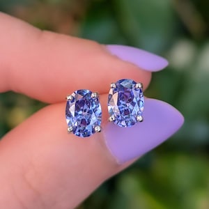 May include: A pair of oval-cut, blue gemstone stud earrings. The earrings have a silver setting with small accent stones around the main gem. The background is blurred, with a focus on the earrings.