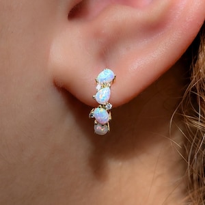 May include: A gold hoop earring featuring a vertical arrangement of four pear-shaped iridescent opals and small clear accent stones. The earring is secured in an earlobe, showcasing the play of color within the white opals and the metallic gold setting.