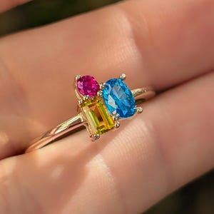 May include: A gold ring featuring a cluster of colorful gemstones. The ring has a pink oval stone, a blue oval stone, and a green rectangular stone. The gemstones are set on a delicate gold band.