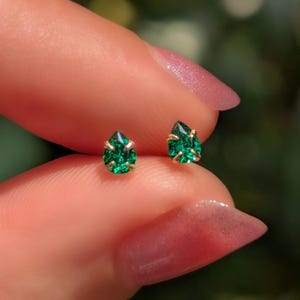 May include: A pair of small, teardrop-shaped emerald green stud earrings with gold-colored settings. The earrings are held between fingers, showcasing their vibrant color and delicate design. The earrings are a jewelry item.