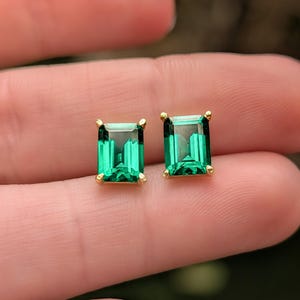 May include: A pair of emerald-cut, green gemstone stud earrings with gold settings. The rectangular earrings display a vibrant, transparent green hue. The gold settings feature small prongs securing the gemstones.