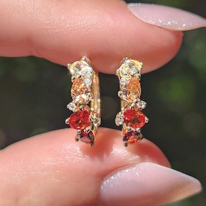 May include: A pair of gold hoop earrings featuring a cluster of multi-colored gemstones in shades of yellow, orange, and red. The stones are arranged in a gradient pattern along the front of each hoop, accented by small, clear, sparkling crystals.